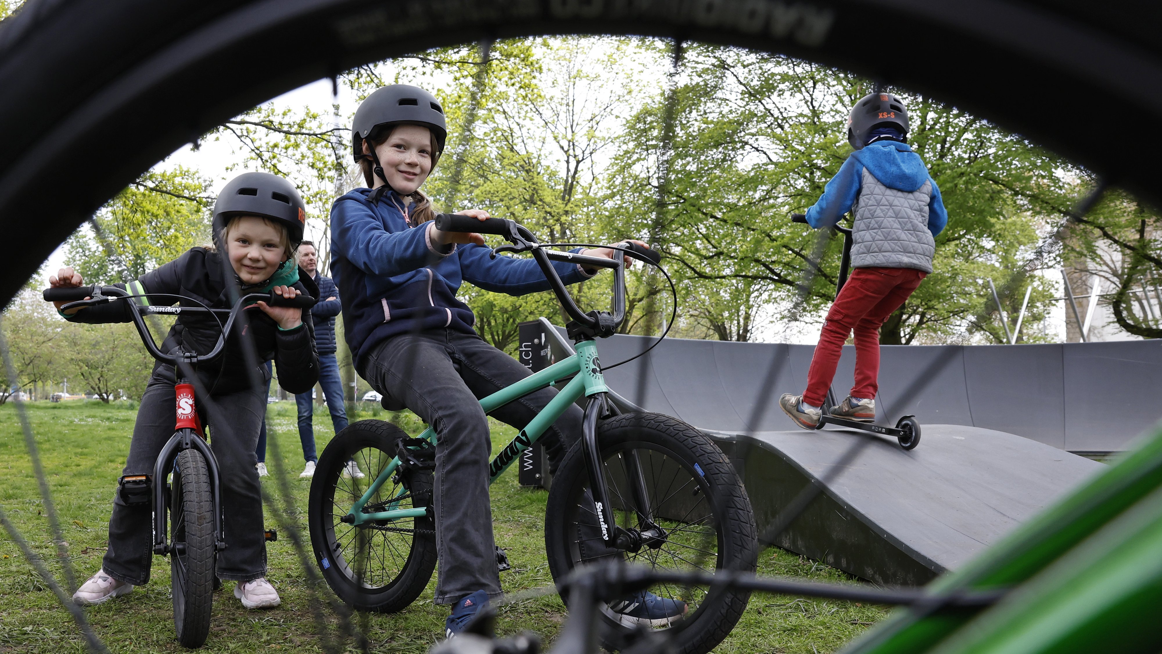 Kinder sitzen auf ihren Fahrrädern neben einem Pumptrackparkour und schauen durch einen Fahrradreifen und dessen Speichen hindurch